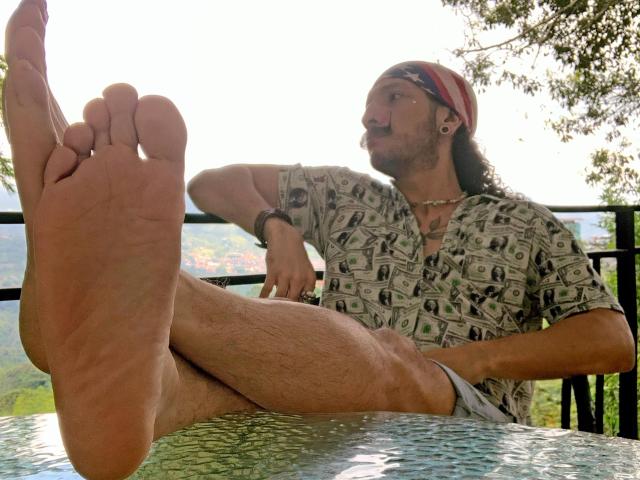 RebelDali69, with a mustache and headband, sits on a metal chair, showing his bare, hairy-assed foot, wearing a money-pattern shirt, and looking to the side.
