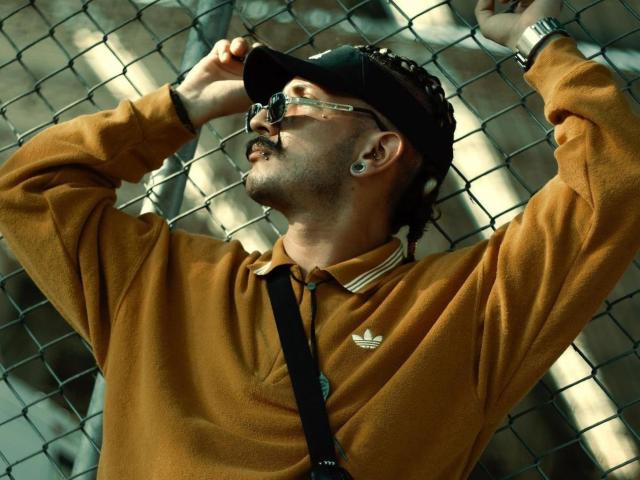 RebelDali69 with glasses, black cap, brown polo, Adidas logo, ear gauges, and black strap, leaning against a chain-link fence, arms raised, sunglasses on, mustache, watch on wrist.