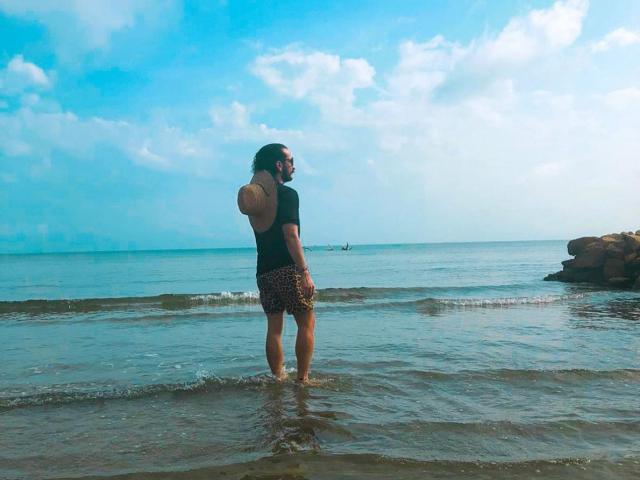 RebelDali69 stands in shallow water, back to camera, wearing a black shirt and leopard print shorts, with wavy brown hair, and a small tattoo on their upper back.