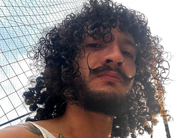 RebelDali69 has curly dark hair, a mustache, and a beard, with a nose ring, looking down at the camera, wearing a white tank top, and a tattoo on his chest.