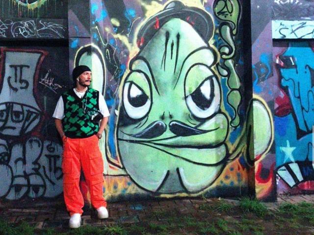 RebelDali69 with green vest, black cap, and orange pants, standing next to a large, graffiti'd green cartoon face with a smug expression, hands in pockets.