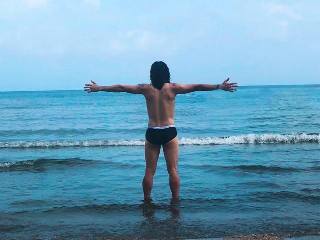 RebelDali69 stands in shallow ocean water, back to camera, arms out, wearing black speedo, showing a toned butt and muscular back, under a blue sky.