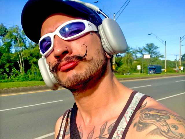 RebelDali69 has a mustache tattoo, white headphones, blue sunglasses, red lips, and a tattooed arm, wearing a tank top and cap, on a sunny road.
