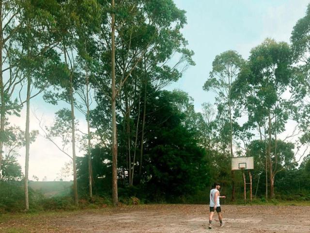 RebelDali69, shirtless with white tank top, black shorts, standing on dusty outdoor basketball court, tall green trees in background, right side of image, holding basketball, daytime.