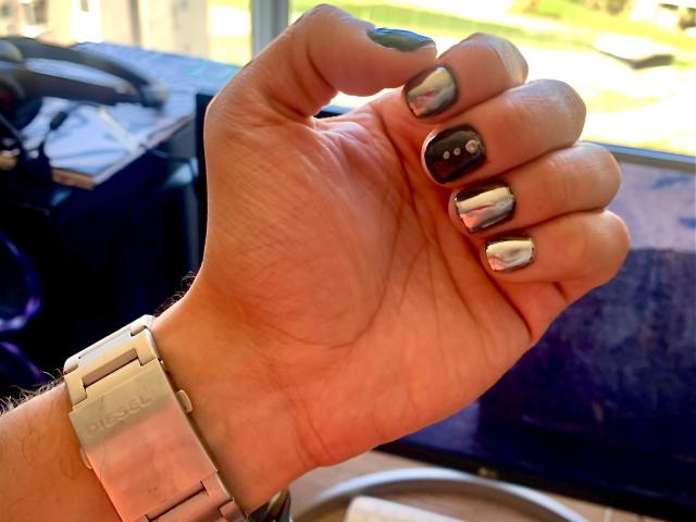 RebelDali69's hand with shiny black and silver nail polish, a silver Rolex watch, and a visible black bag in the blurry background.