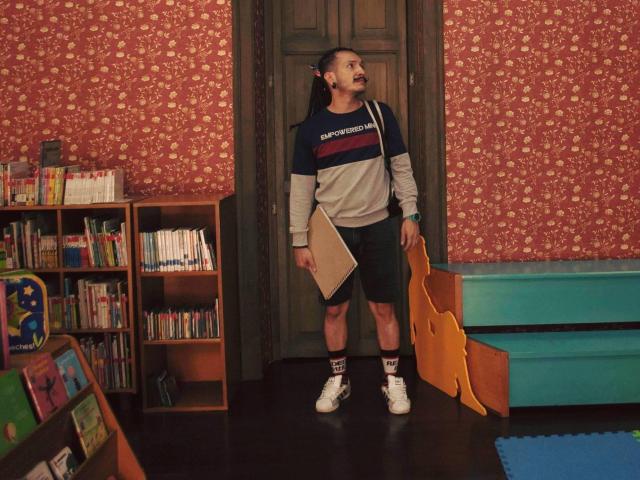 RebelDali69 stands in a room with red floral wallpaper, wearing a sports shirt, black shorts, white socks, and sneakers, holding a book, with a wooden door behind.