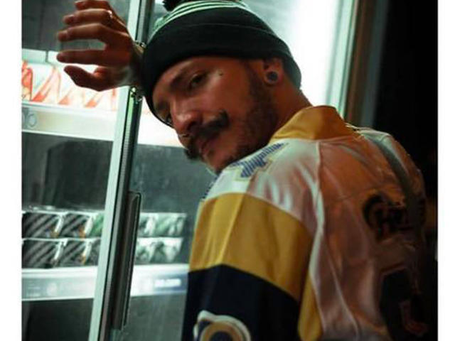 RebelDali69 with a mustache, wearing a black beanie and a yellow, white, and blue hockey jersey, standing in front of a glass freezer. His ear is stretched.