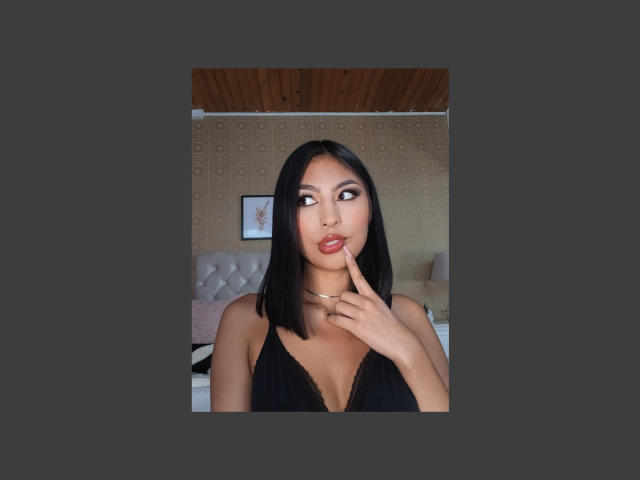 CarlaRusso with straight black hair, pouty lips, and a finger on her chin, wearing a black lace top, showing her tits, in a bedroom with a wooden ceiling.