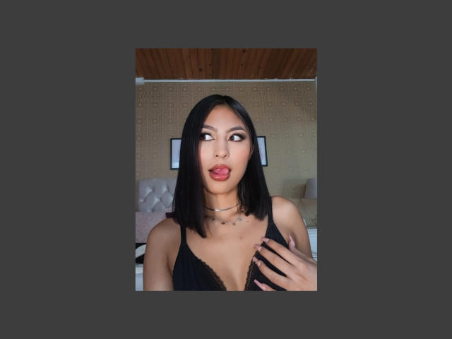 CarlaRusso with black bob, pouty red lips, looking sideways, black lace bra, small tits, hand on chest, wooden ceiling, beige wall, flat-screen TV in background, dark border.