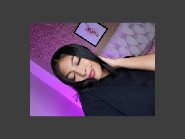 CarlaRusso with closed eyes, black hair, and a slight smile, wearing a black shirt, her hand touching her neck, in a purple-lit room with a tufted pillow.