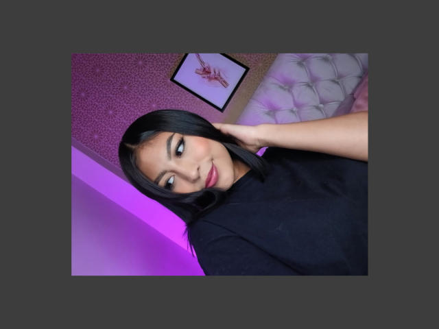 CarlaRusso with black hair, smiling, wearing a black top, hand on her head, purple light, tufted white pillow, framed artwork in the background, looking at the camera.