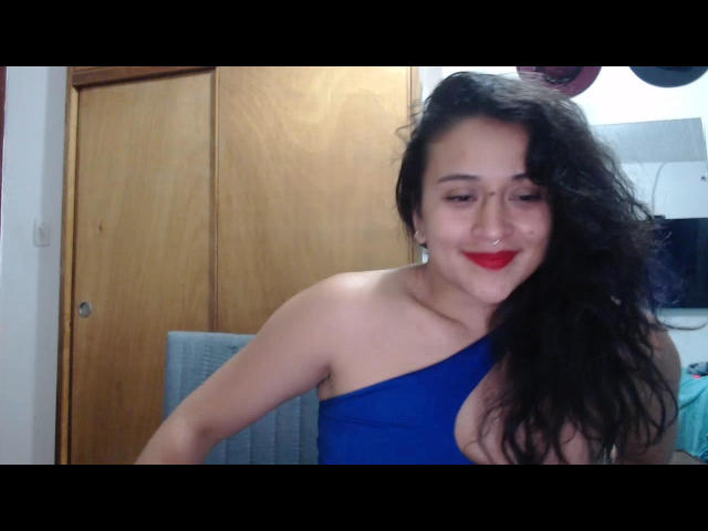 GiovannaBellaC with long black hair, red lipstick, and a blue one-shoulder top, smiling at the camera, showing her tits. She's got a nose ring and is sitting.