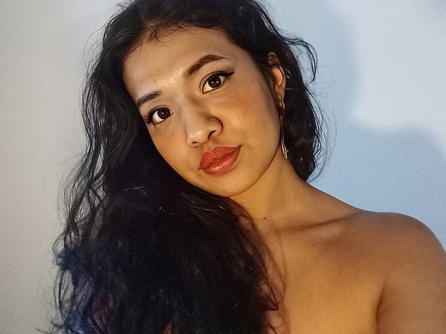 LucyMorga with long, wavy black hair, brown eyes, and glossy red lips, topless, showing her bare shoulders and small, perky tits. Her skin is smooth and she's smiling.