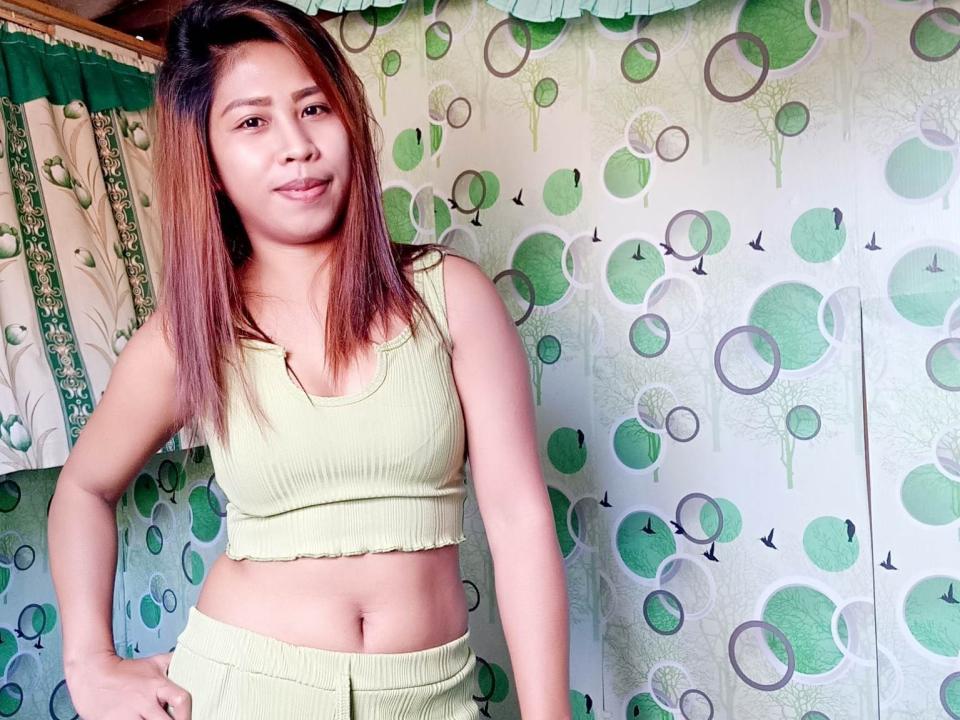 LuksapGaw trägt ein hellgrünes, geripptes Crop-Top und einen passenden Rock, wobei ihr flacher Bauch zu sehen ist. Sie hat braunes Haar mit rötlichen Strähnen und ein leichtes Lächeln, und sie steht selbstbewusst da..