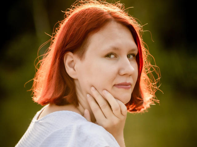 Suniabdsm-hot has red, wavy hair, fair skin, and a slight smile. She's touching her cheek with her right hand, wearing a white top. Soft sunlight highlights her face.