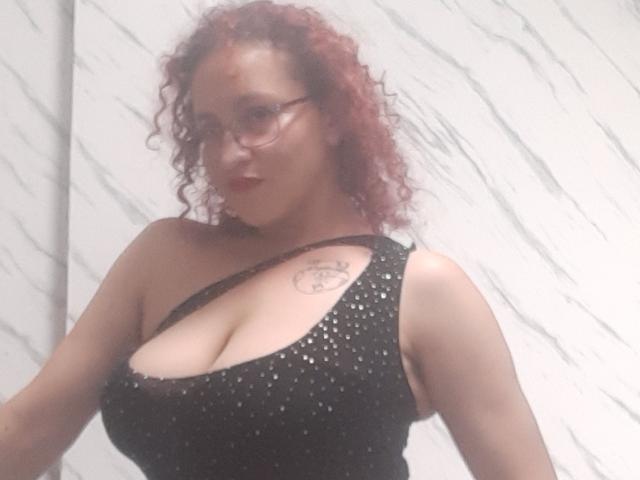 CrystalBush with curly red hair, glasses, and black sequined dress, showing big boobs and a tattoo on her right chest, standing against a white marble wall.