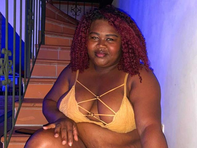 BritannyJhonson, dark-skinned with red curly hair, wearing a yellow lace bra with criss-cross straps, sitting on stairs, smiling, showing her big boobs, and holding her knee.