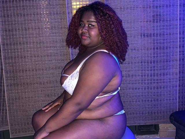 BritannyJhonson, a curvy dark-skinned woman with red curly hair, wearing a white lace bra and matching thong, sits on a surface, showing her large ass and boobs.