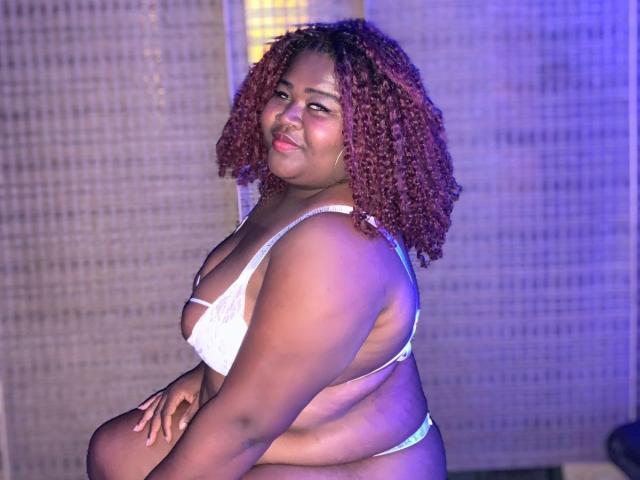 BritannyJhonson with curly red hair, white bra, and thong, showing her big ass and big boobs, smiling at the camera, sitting down, with a blue and yellow light.