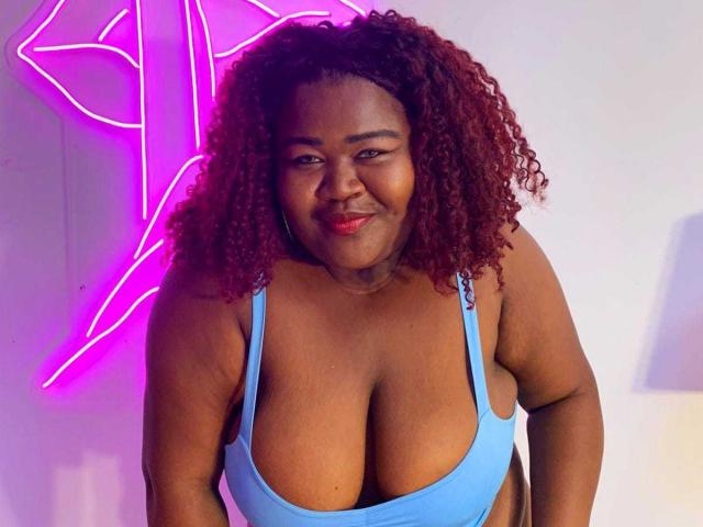 BritannyJhonson with curly red hair, dark skin, and large tits in a blue sports bra, smiling at the camera. Neon pink light in background.