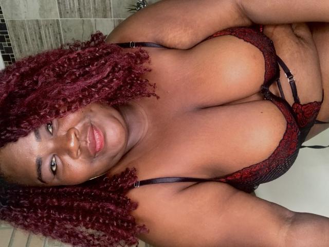 BritannyJhonson with curly red hair, wearing red and black lace bra, showing her large tits and smiling at the camera, in a bathroom. Her dark brown skin is smooth.