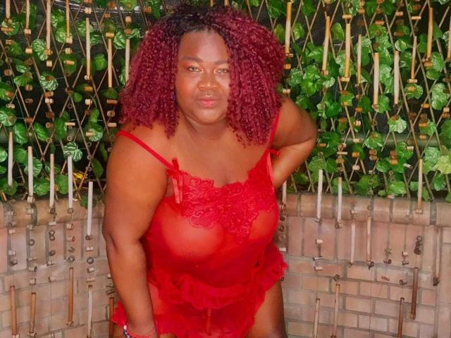 BritannyJhonson with red curly hair, wearing sheer red lace lingerie, exposing her tits, standing with a confident smile, hand on hip.