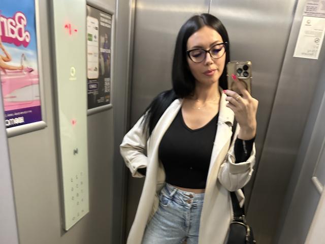Nadeen with black hair, glasses, white jacket, black top, and blue jeans, holding phone, taking mirror selfie in elevator, showing her tits in tight top.