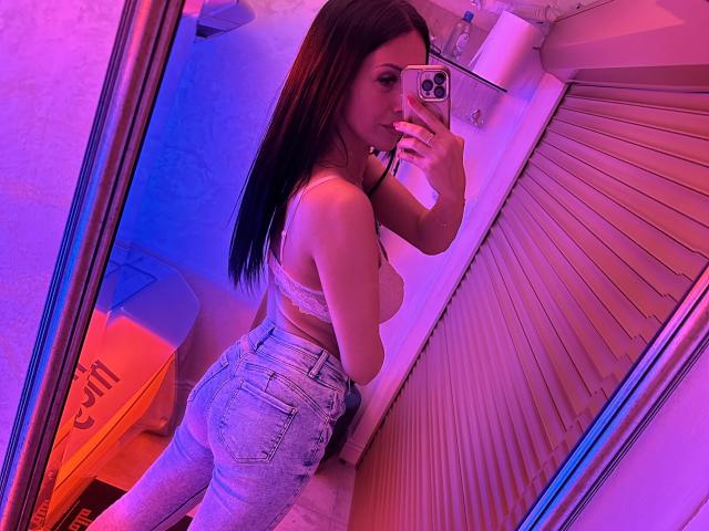 Nadeen mit langen braunen Haaren, die einen weißen Spitzen-BH und hochgeschnittene blaue Jeans trägt, macht ein Selfie vor einem Spiegel und zeigt dabei ihren Hintern und ihre kleinen Brüste, bei violetter und blauer Beleuchtung..