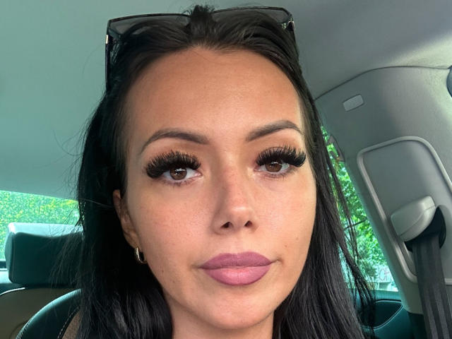 Nadeen has dark brown eyes, long black eyelashes, pink lipstick, and is wearing black sunglasses on her head. Her tan skin is visible in the car's interior.