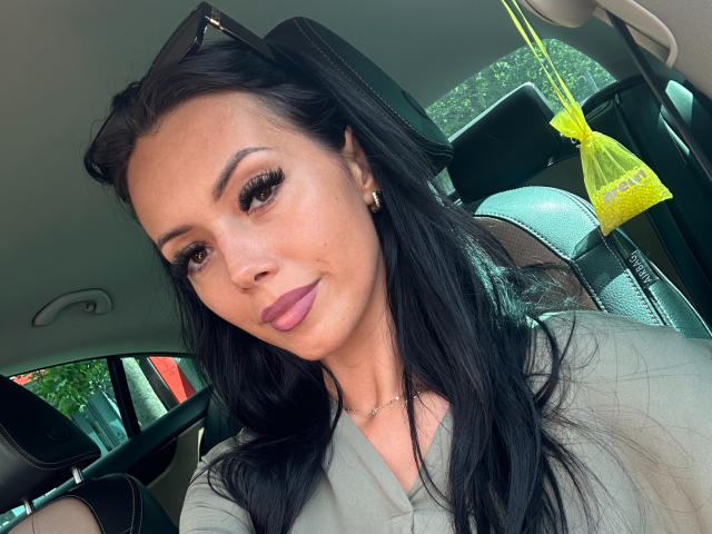 Nadeen with long black hair, wearing a grey shirt, has big ass, brown eyes, thick eyelashes, pink lipstick, and a necklace. She's in a car with yellow bag.