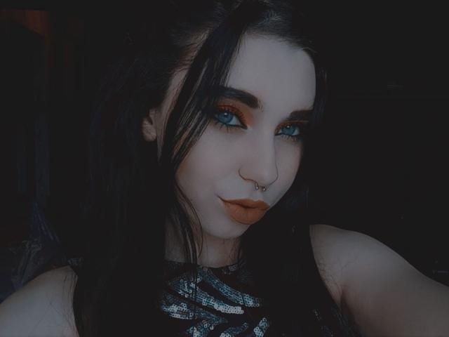 LegendBrenda has pale skin, blue eyes, dark makeup, orange lipstick, black hair, and a nose ring. She's wearing a shiny black top, looking sexy in a dark setting.