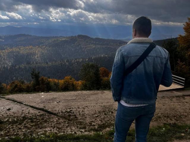 GordonDesire è di spalle rispetto alla telecamera, indossa una giacca di jeans blu e pantaloni jeans, con una cintura nera attraversata sul petto, e guarda un paesaggio montuoso avvolto nella nebbia..