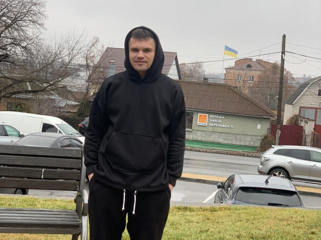 GordonDesire stands in front of a wooden bench, wearing a black hoodie and black pants, with hands in pockets, looking slightly to the side with a subtle smile.
