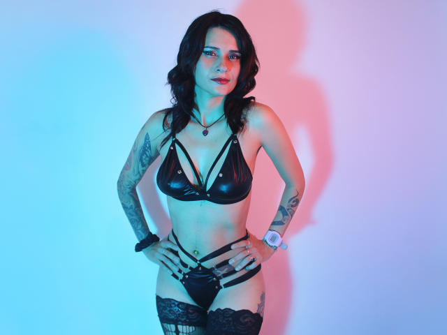 HannieJones with dark wavy hair, black leather bra, strappy black panties, tattoos on arms, lace thigh-highs, hands on hips, blue and pink lighting, confident expression.