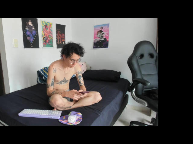 BlakeTyler, senza maglietta e con tatuaggi, è seduto su un letto nero, con le gambe incrociate, e guarda un telefono. Una tastiera e un mouse sono sul letto. Una sedia da gaming grigia è nelle vicinanze..