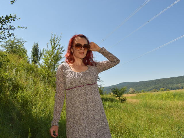 KateRouge with red hair, wearing a speckled dress, brown sunglasses, standing in a green field, hand on head, smiling, sunny day, clear blue sky, power lines in background.