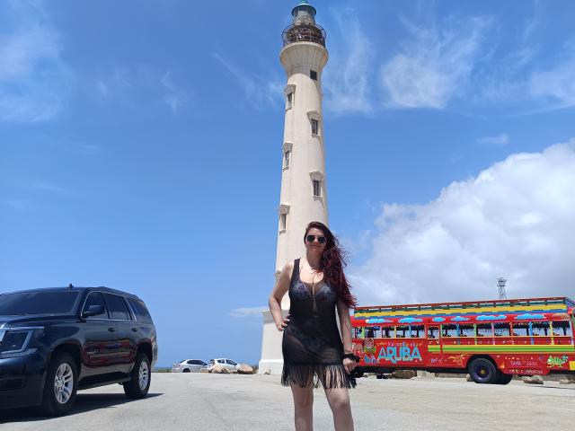 Katherin, una mujer de origen latino con cabello rojo largo, un vestido negro y gafas de sol, está de pie frente a un alto faro blanco, con un autobús turístico colorido y un SUV negro a la izquierda..