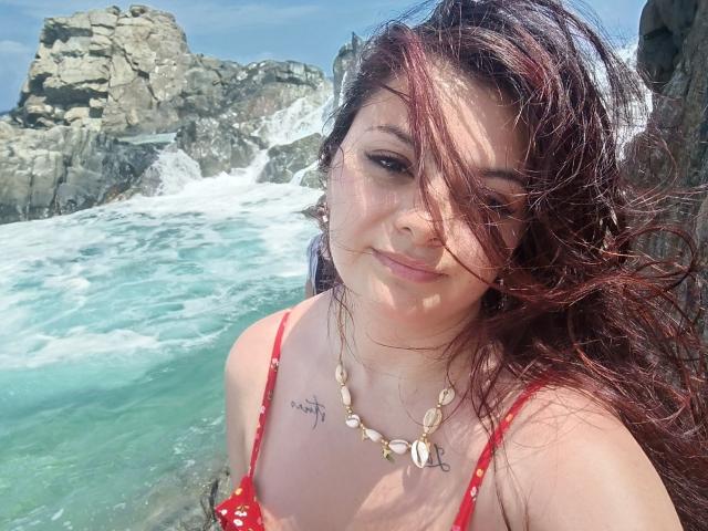 Katherin Latin tiene el pelo rojo, un tatuaje en el pecho derecho y lleva un bikini rojo con un collar de conchas. Sus pechos son pequeños y está sonriendo a la cámara..