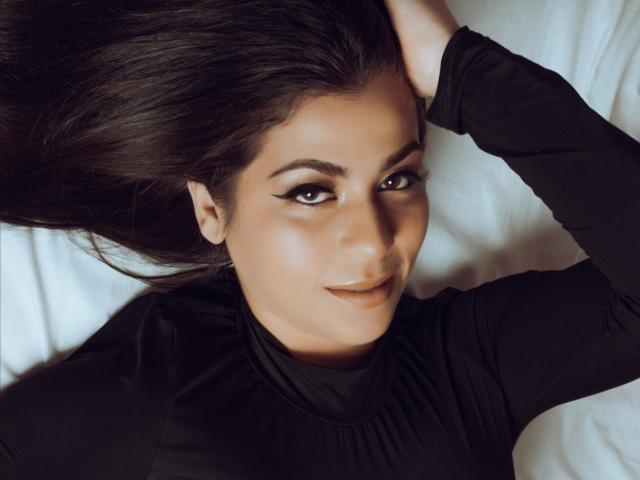 BelleeRose with dark, flowing hair, black eyeliner, and a subtle smile, wearing a black top, lying on a white bed, hand in her hair, looking at the camera.
