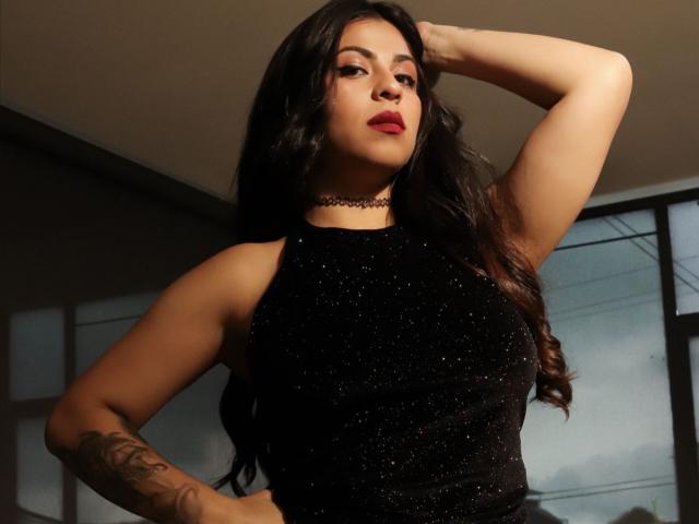 BelleeRose with dark wavy hair, wearing a sparkly black sleeveless top, choker, hand on head, tattoo on arm, red lipstick, confident expression, standing in dimly lit room.