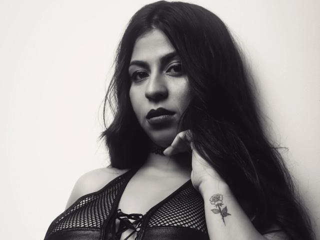Black-and-white photo of BelleeRose with long dark hair, wearing a mesh halter top, showing her tits, hand on chin, rose tattoo on right wrist, serious expression.