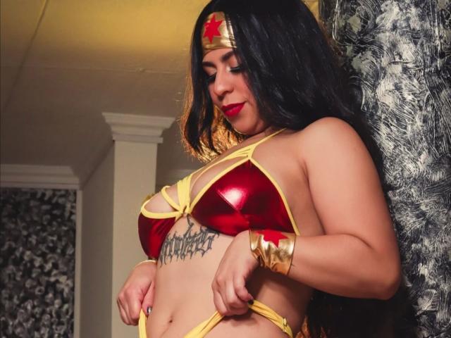 BelleeRose with long black hair, red shiny bra, yellow straps, gold headband, and gold bracelet, showing off her large tits and tattooed belly, looking down with a smile.