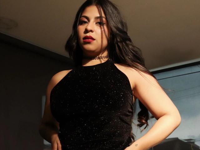 BelleeRose with long black hair, wearing a sparkly black halter top, red lipstick, and a choker, standing with hands on hips, looking confident, showcasing her big tits.