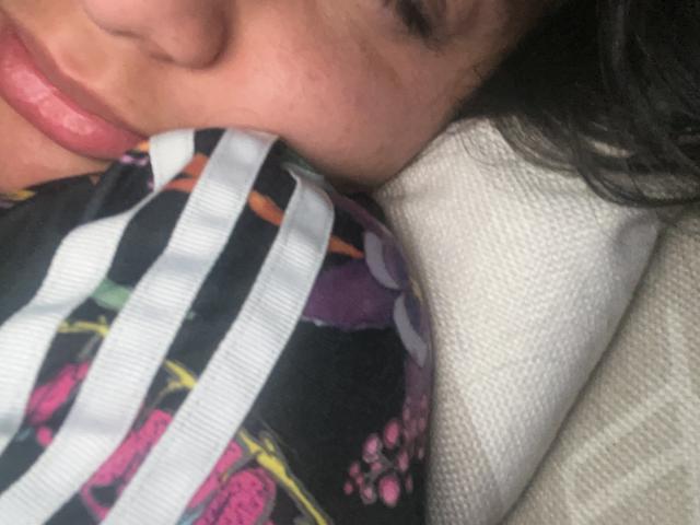 El rostro de DianaFernandezz es parcialmente visible, mordiendo un tejido colorido con rayas negras y blancas, flores rosas y patrones morados, con la cabeza apoyada en un cojín blanco..