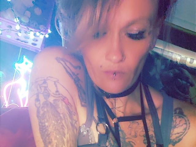 RenegadeVyx with tattoos on arms, wearing black harness, nose ring, and lip piercing, has short blonde hair, and is looking down with a slightly parted mouth.