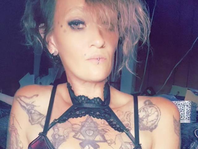 RenegadeVyx with pink-dyed hair, black lace top, multiple tattoos on chest and shoulders, nose piercing, and lip piercing, looking at camera with a slight smirk.