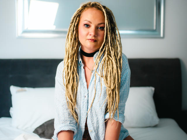 KayaBlonde mit blonden Dreadlocks, trägt ein blau kariertes Hemd und eine schwarze Halskette, sitzt auf einem Bett mit weißen Kissen und blickt leicht zur Seite, mit einem ernsten Gesichtsausdruck..