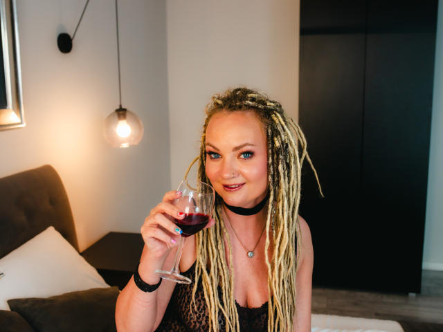 KayaBlonde, avec des tresses blondes, tenant un verre de vin rouge, portant un collier noir et un haut en dentelle, souriante, exhibant sa poitrine, dans une pièce confortable..