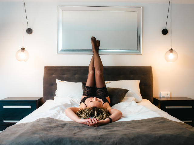 KayaBlonde est allongée sur un lit, les jambes en l'air, vêtue de lingerie en dentelle noire et de bas hauts, avec ses cheveux blonds éparpillés, dans une pose sexy et détendue..