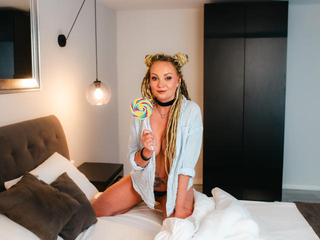 KayaBlonde with blonde braids, holding a colorful swirl lollipop, wearing an open check shirt, sitting on a bed, showing a tattoo on her right hip, smiling.